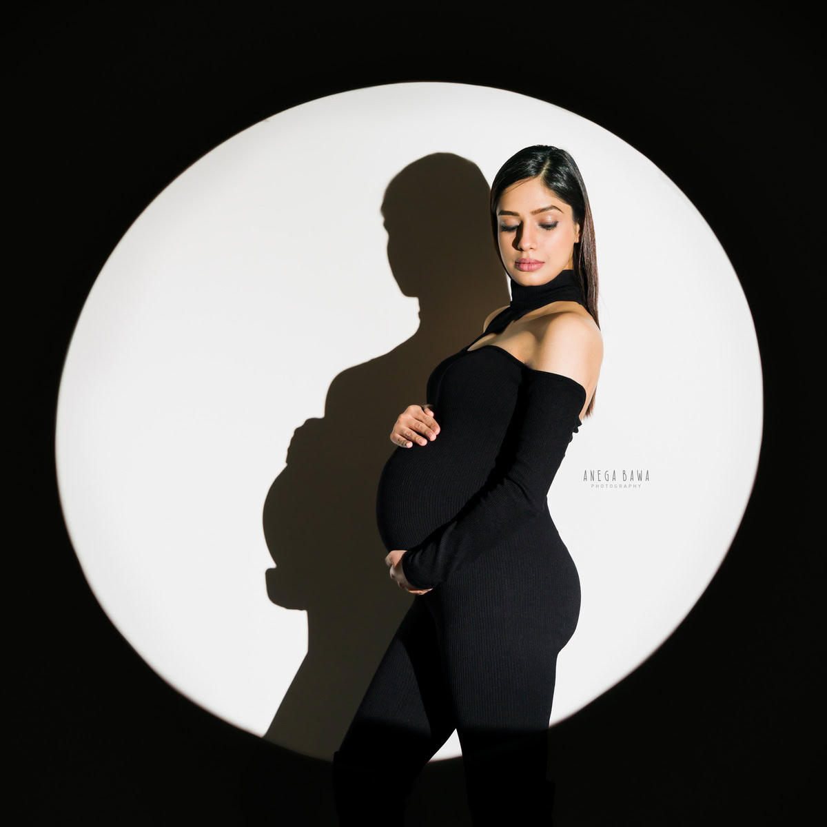 Elegant maternity photo session in Delhi featuring a woman in a black jumpsuit against a white backdrop, highlighted by artistic lighting. Captured by Anega Bawa Photography, Gurgaon