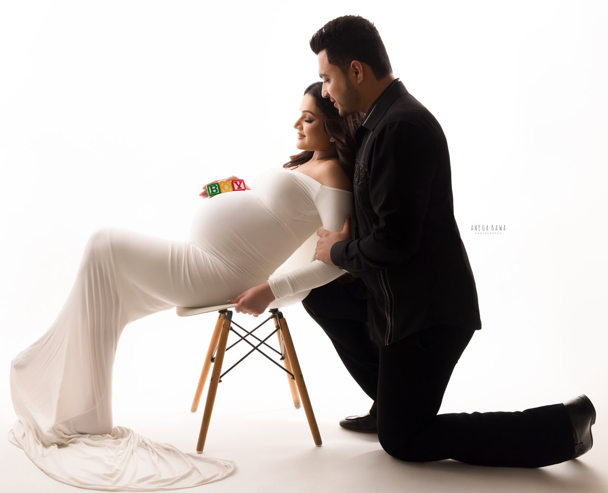 Pregnant woman in white dress with partner holding her belly with baby blocks, maternity photoshoot, studio setting, Anega Bawa Photography, timeless pregnancy portrait, family love and anticipation.