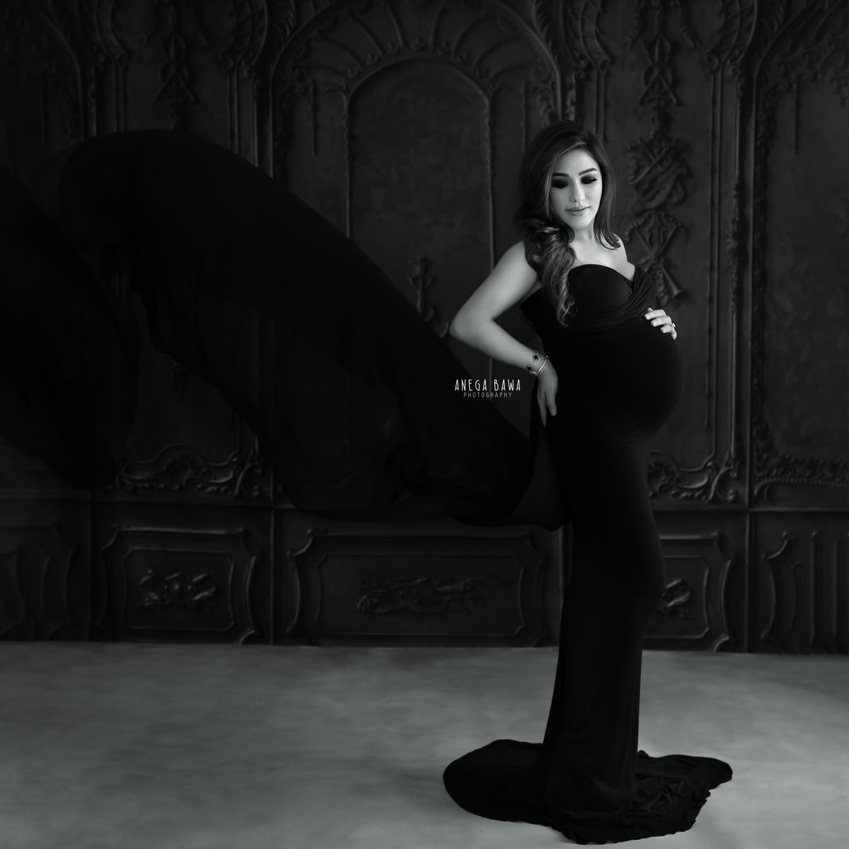 Elegant pregnant woman portrait in black and white, maternity photography by Anega Bawa, featuring a stylish gown and a classy indoor setting.