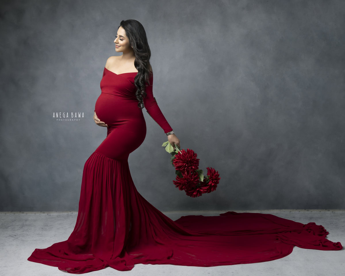 Elegant pregnant woman in red gown holding red flowers in a studio photoshoot.