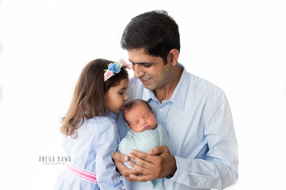 Newborn Photography in Delhi: A baby wrapped in blue with father and sibling on a white backdrop. Photographer: Anega Bawa Photography, Gurgaon.