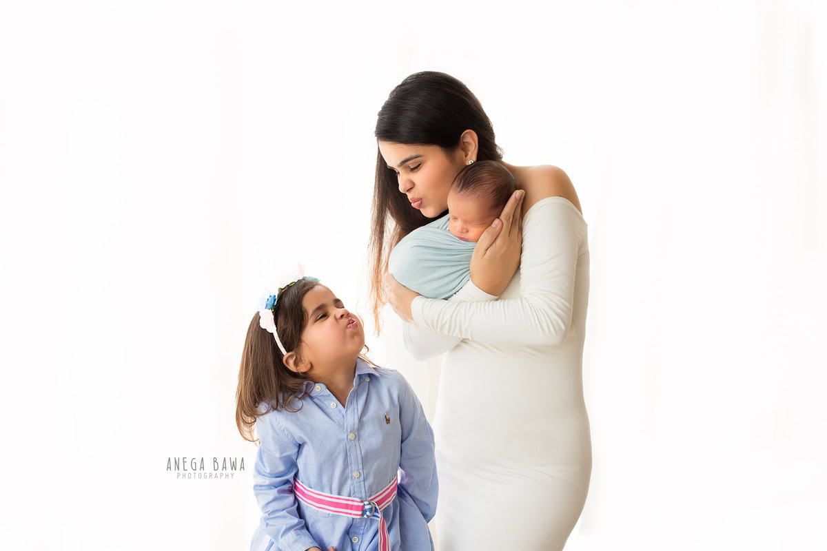 Newborn Photography in Delhi: A baby wrapped in blue with mom and sibling on a white backdrop. Photographer: Anega Bawa Photography, Gurgaon.