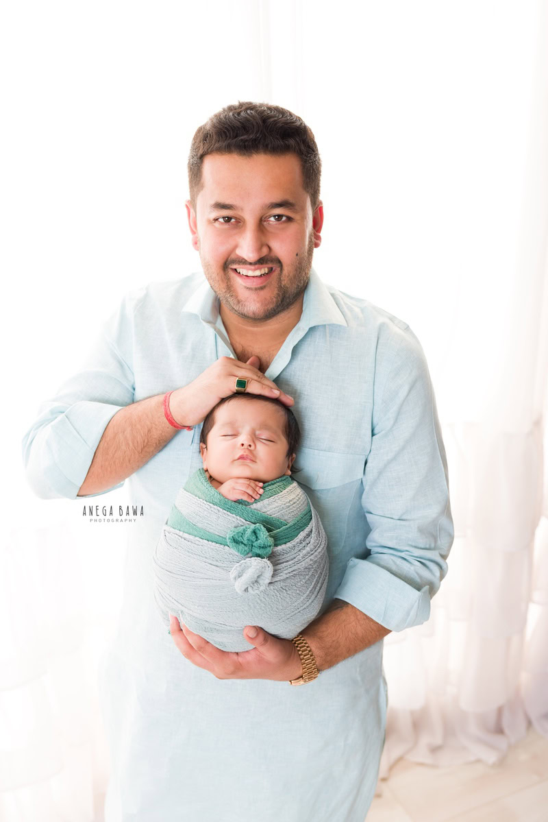 Newborn Photography in Delhi: Father holding newborn wrapped in grey-green fabric against a white backdrop. Photographer: Anega Bawa Photography, Gurgaon.