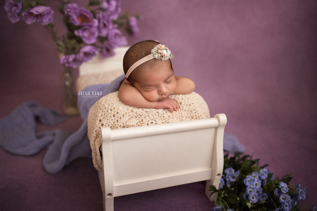 Newborn Photography in Delhi: Off-White Cot, Lavender Backdrop, Lavender Blue Flowers. Photographer: Anega Bawa Photoshoot, Gurgaon Noida.