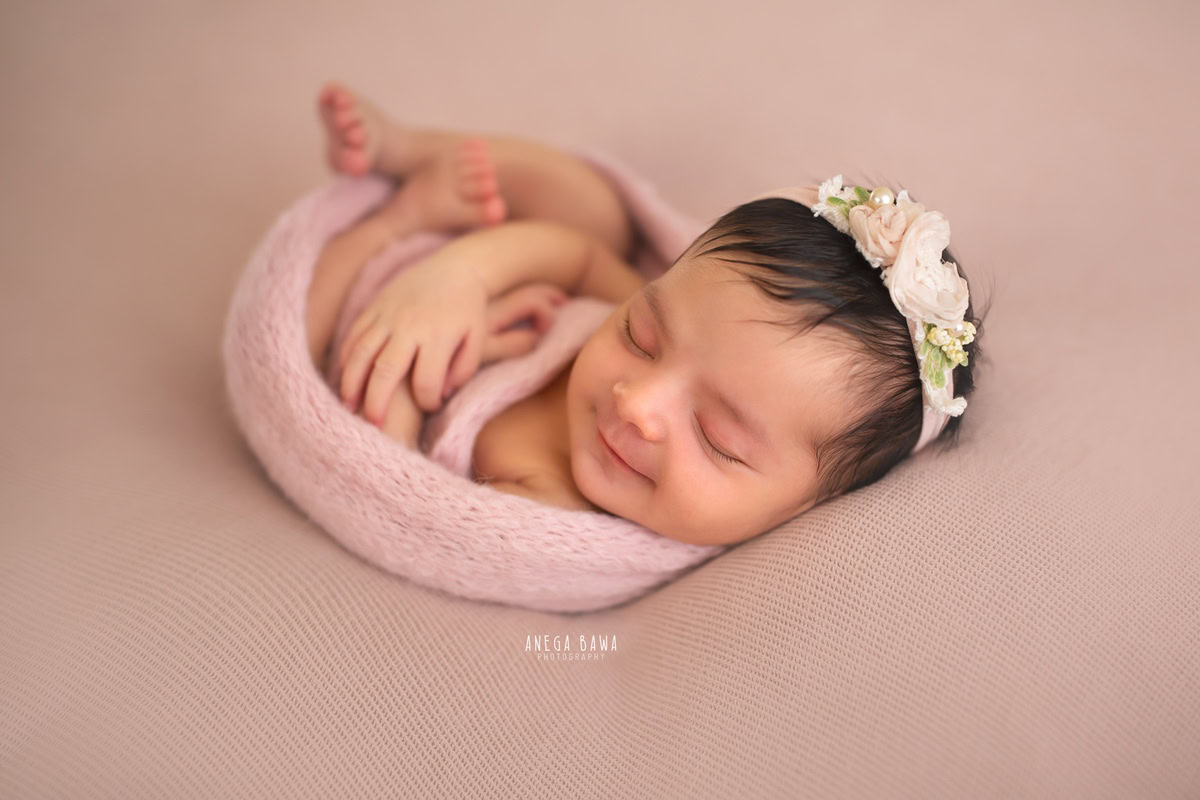Newborn Photography in Delhi: Cute Pose with Pink Wrap and Floral Headband on Beige Backdrop. Photographer: Anega Bawa, Gurgaon Noida.