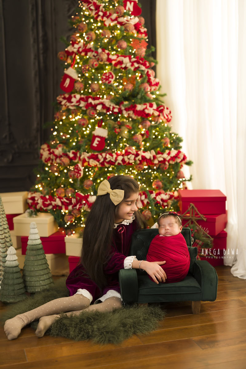 Newborn Photography in Delhi: Red Wrap with Sibling, Green Rug, and Xmas Tree. Photographer: Anega Bawa, Gurgaon Noida.