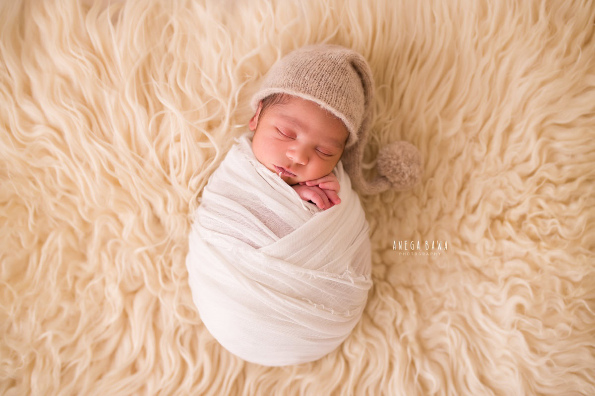 Newborn Photography in Delhi: White Wrap with Brown Cap on Beige Rug. Photographer: Anega Bawa, Gurgaon Noida.