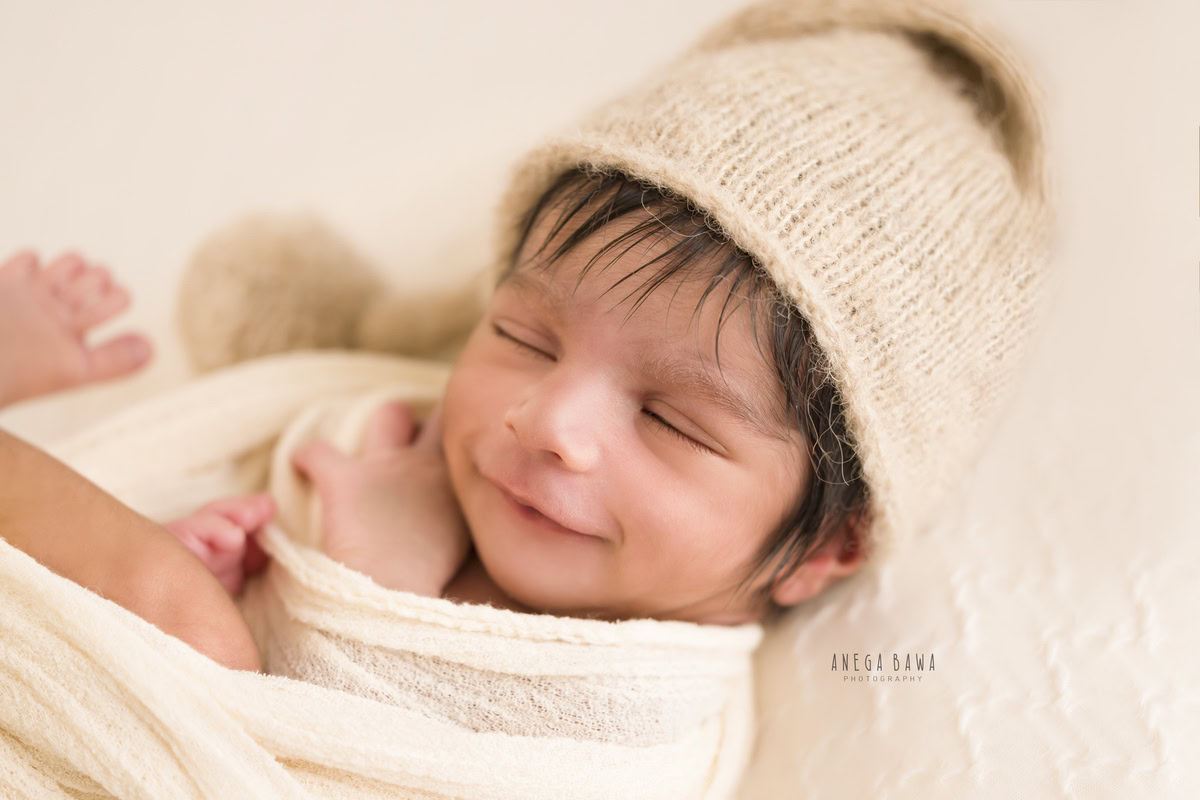 Newborn Photography in Delhi: White Wrap, Brown Cap, Cute Smiling Pose. Photographer: Anega Bawa, Gurgaon Noida.