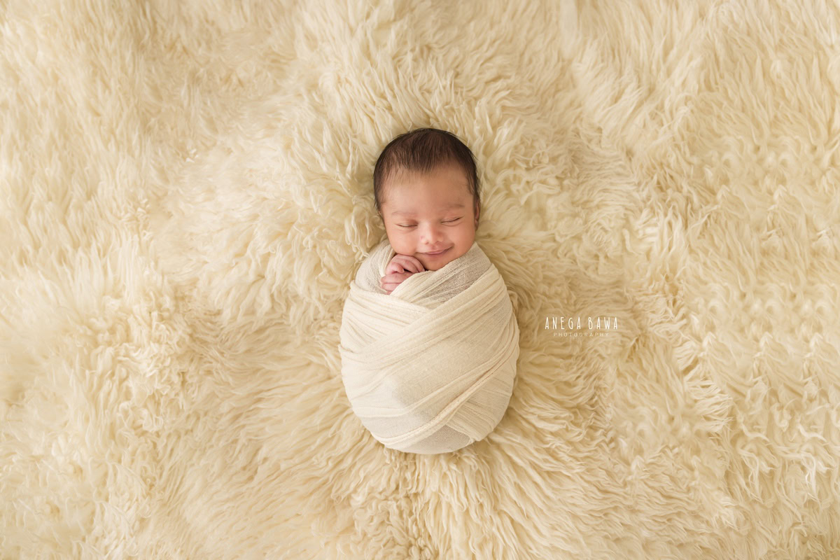 Newborn Photography in Delhi: White Wrap, Cute Smiling Pose, Beige Rug. Photographer: Anega Bawa, Gurgaon Noida.
