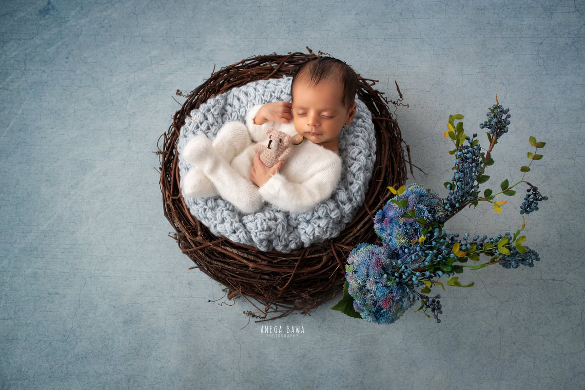 Newborn Photography in Delhi: White Baby Suit, Blue Backdrop with Blue Bushes. Photographer: Anega Bawa, Gurgaon Noida.