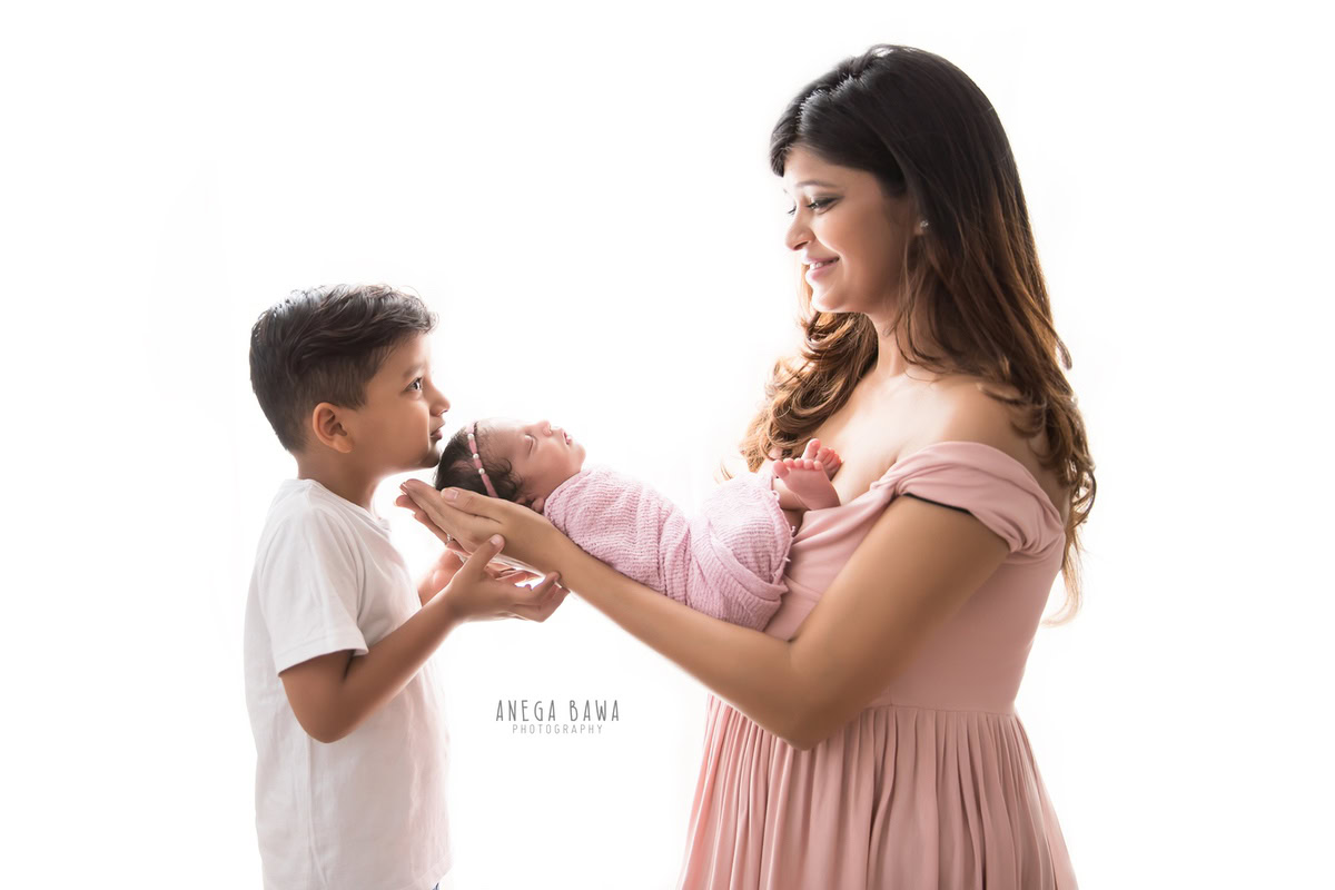 Newborn Photography in Gurgaon: Mother holding newborn with sibling, wrapped in pink, against a white backdrop. Photographer: Anega Bawa, Delhi.