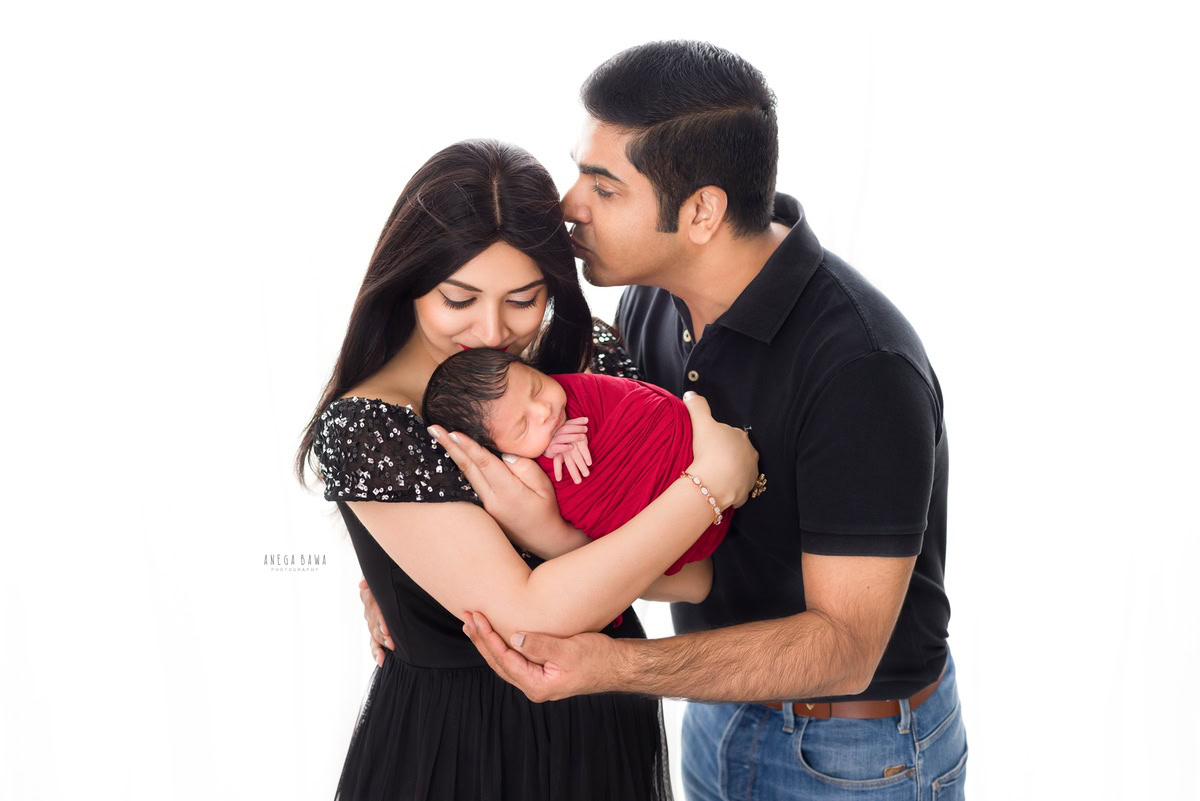 Newborn Photography in Gurgaon: Family kissing pose with newborn wrapped in red, against a white backdrop. Photographer: Anega Bawa, Delhi.