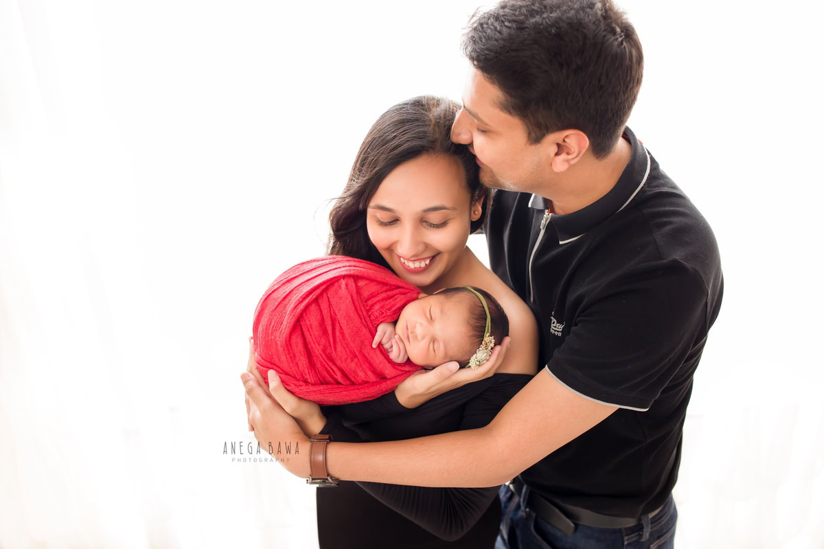 Newborn Photography in Gurgaon: Family with newborn wrapped in red against a white backdrop. Photographer: Anega Bawa, Delhi.
