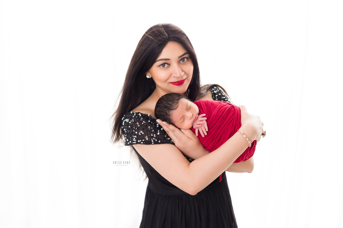 Newborn Photography in Gurgaon: Mother holding newborn wrapped in red against a white backdrop. Photographer: Anega Bawa, Delhi.
