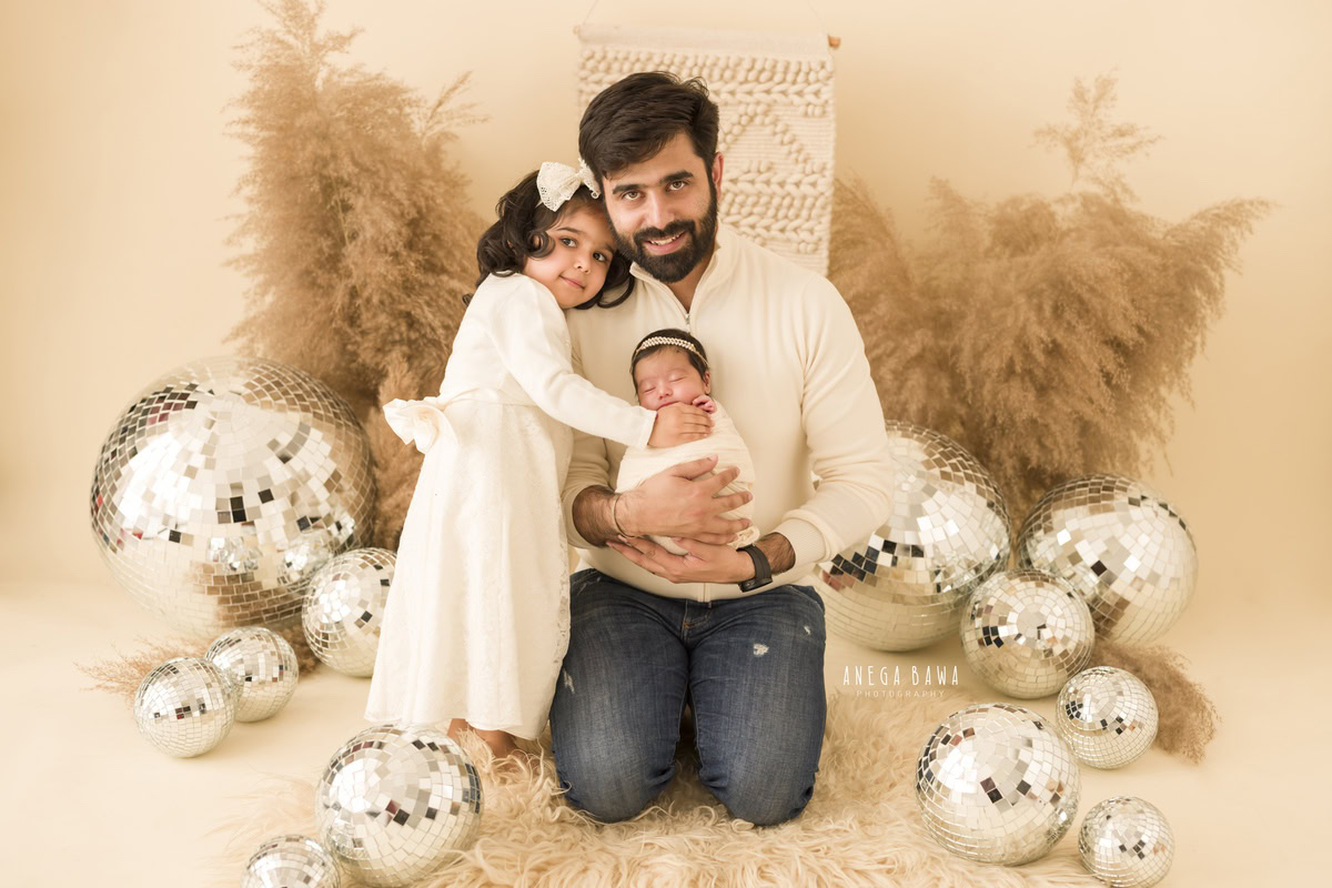 Newborn Photography in Gurgaon: Family portrait with newborn wrapped in white, featuring a disco ball and pampas grass against a beige backdrop. Photographer: Anega Bawa, Delhi.