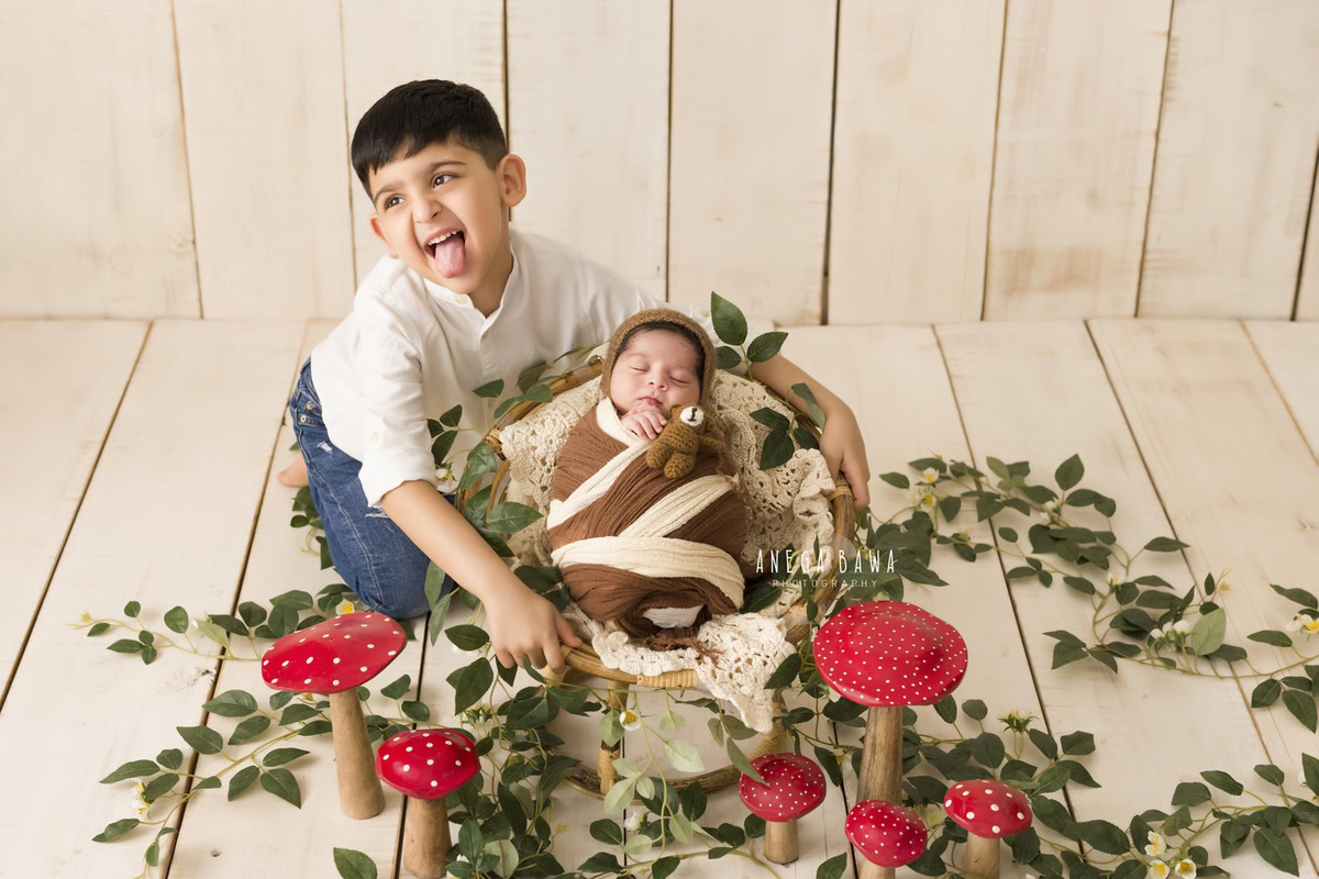 Newborn Photoshoot in Delhi: Beige and Brown Wrap, Sibling Pose on Wooden Cot with Red and White Mushroom Plant, Leaves on Floor, Beige Backdrop. Photographer: Anega Bawa Photography, Gurgaon Noida.
