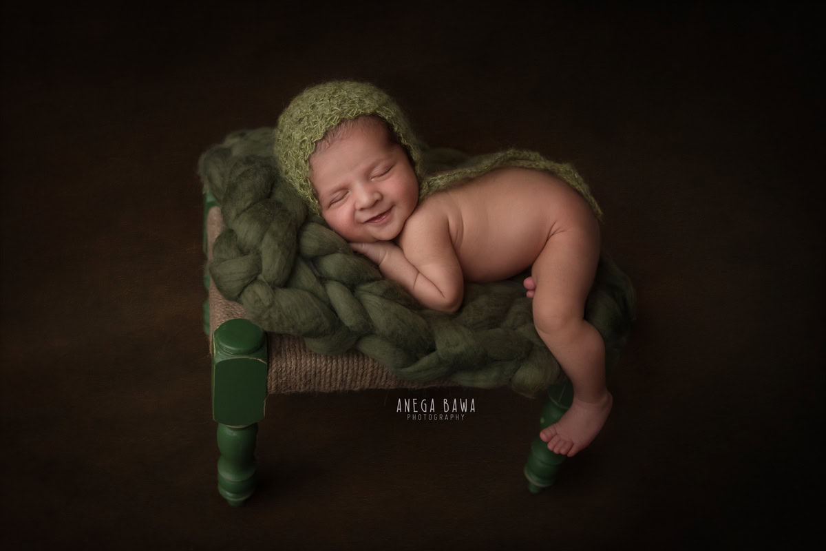 Newborn Photoshoot in Delhi: Green Cap, Wooden Stool, Cute Smiling Pose, Black Backdrop. Photographer: Anega Bawa Photography, Gurgaon Noida.