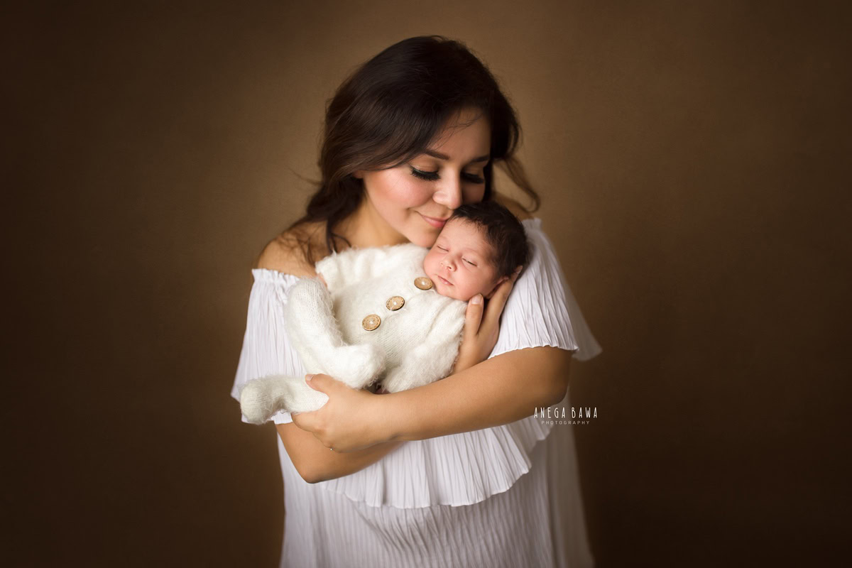 Newborn Photoshoot in Delhi: Mom with her newborn in a white babysuit against a brown backdrop. Photographer: Anega Bawa Photography, Gurgaon.