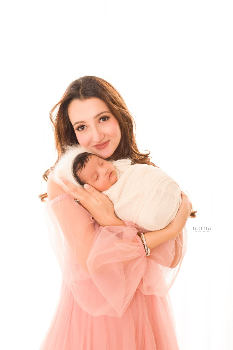 Newborn Photoshoot in Delhi: Mom holding baby in a white wrap and white cap against a white backdrop. Photographer: Anega Bawa Photography, Gurgaon.