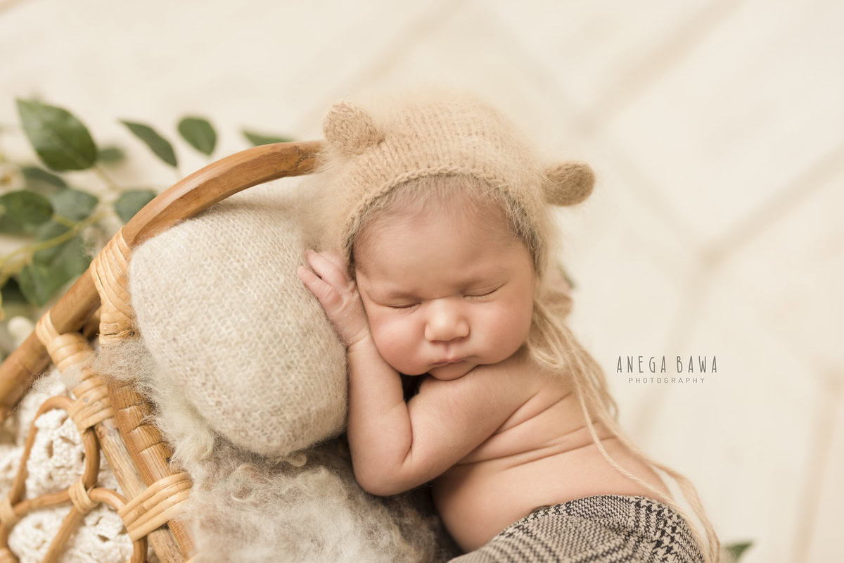 Newborn Photoshoot in Delhi: Sleeping Pose with Brown Cap, Green Leaves, and White Backdrop. Photographer: Anega Bawa Photography, Gurgaon Noida.
