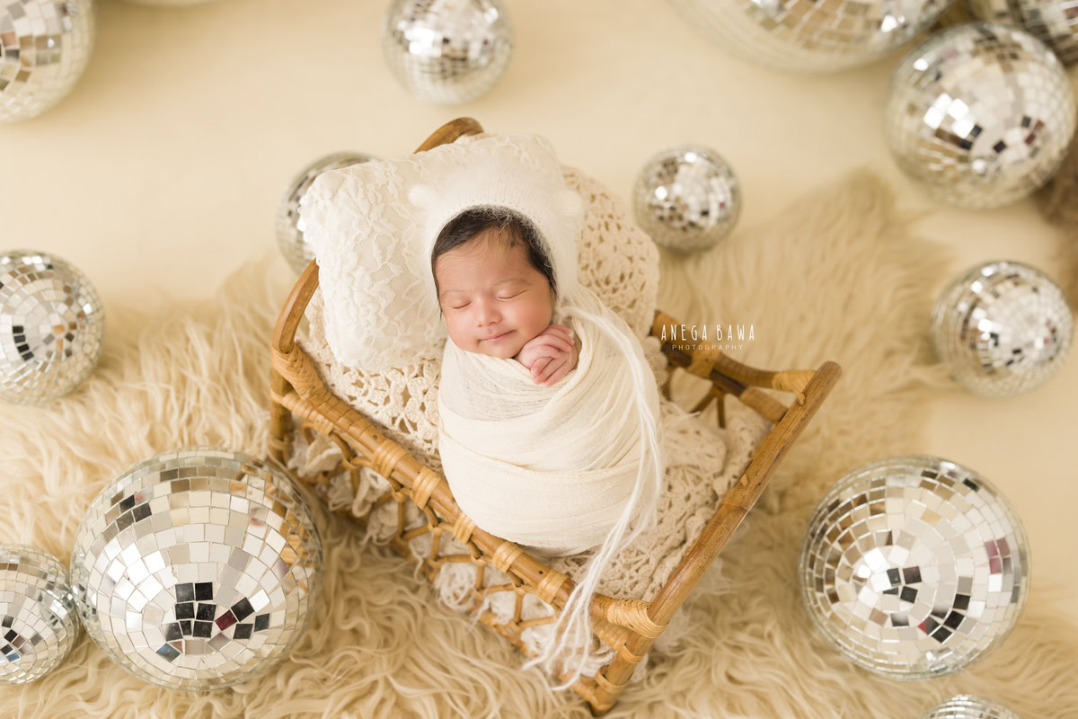 Newborn Photoshoot in Delhi: A baby wrapped in white with a white cap lying on a beige rug under a disco ball against a beige backdrop. Photographer: Anega Bawa Photography, Gurgaon Noida.