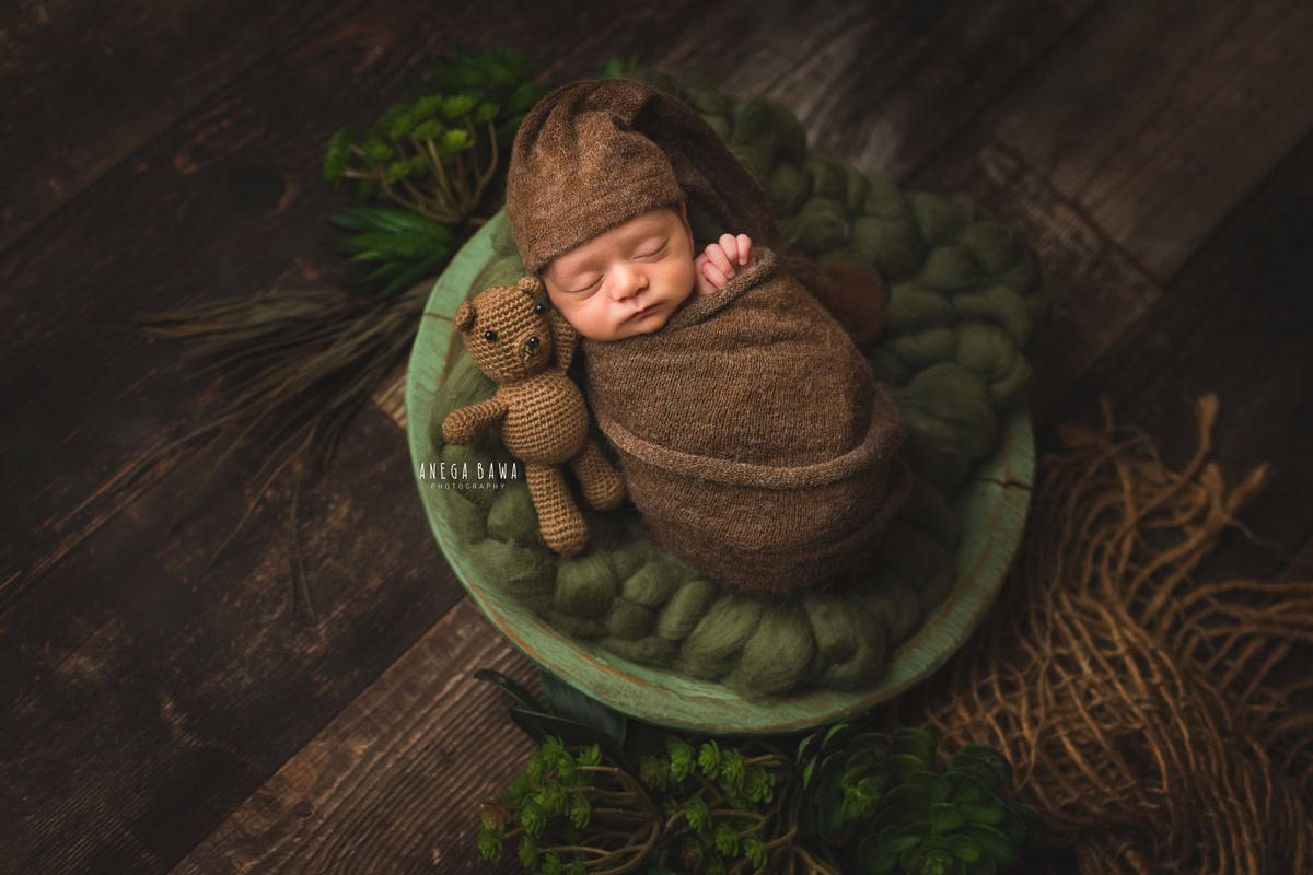 Newborn Photoshoot in Gurgaon: Green Basket with Brown Wrap, featuring Stuff Toy and Brown Cap on Wooden Floor. Photographer: Anega Bawa, Delhi Noida.