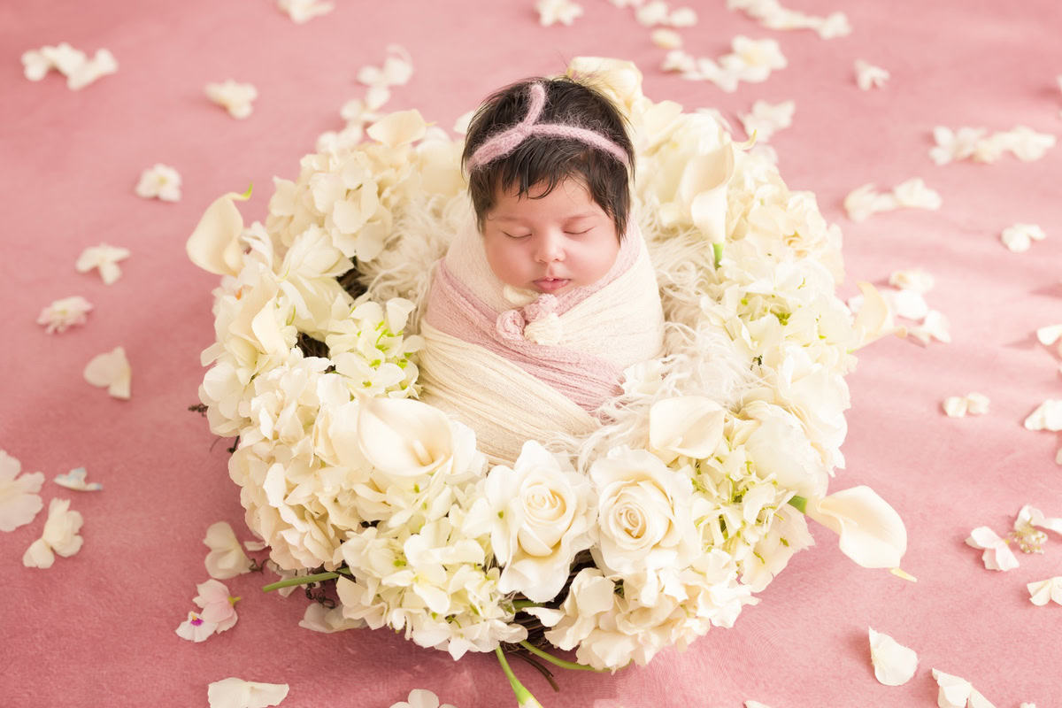 Newborn Photoshoot in Gurgaon: A baby lying on a pink rug wrapped in a pink and white wrap with white petals on the floor and a floral basket nearby. Photographer: Anega Bawa Photography, Delhi Noida.