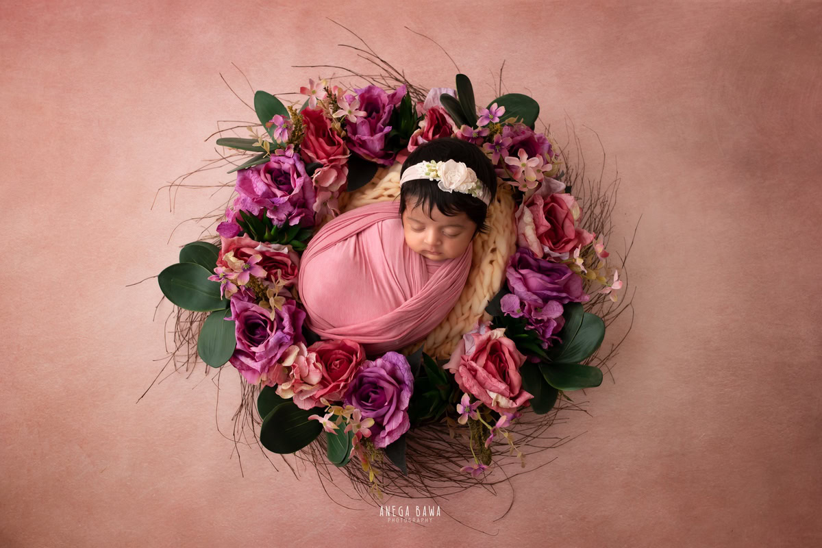 Newborn Photoshoot in Gurgaon: Pink Wrap with Floral Basket on Pink Backdrop. Adorable headband adds charm. Photographer: Anega Bawa Photography, Delhi Noida.