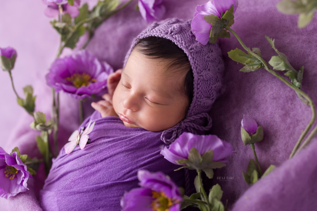 Newborn Photoshoot in Gurgaon: Adorable baby wrapped in purple surrounded by purple flowers against a lavender backdrop. Photographer: Anega Bawa Photography, Delhi Noida.