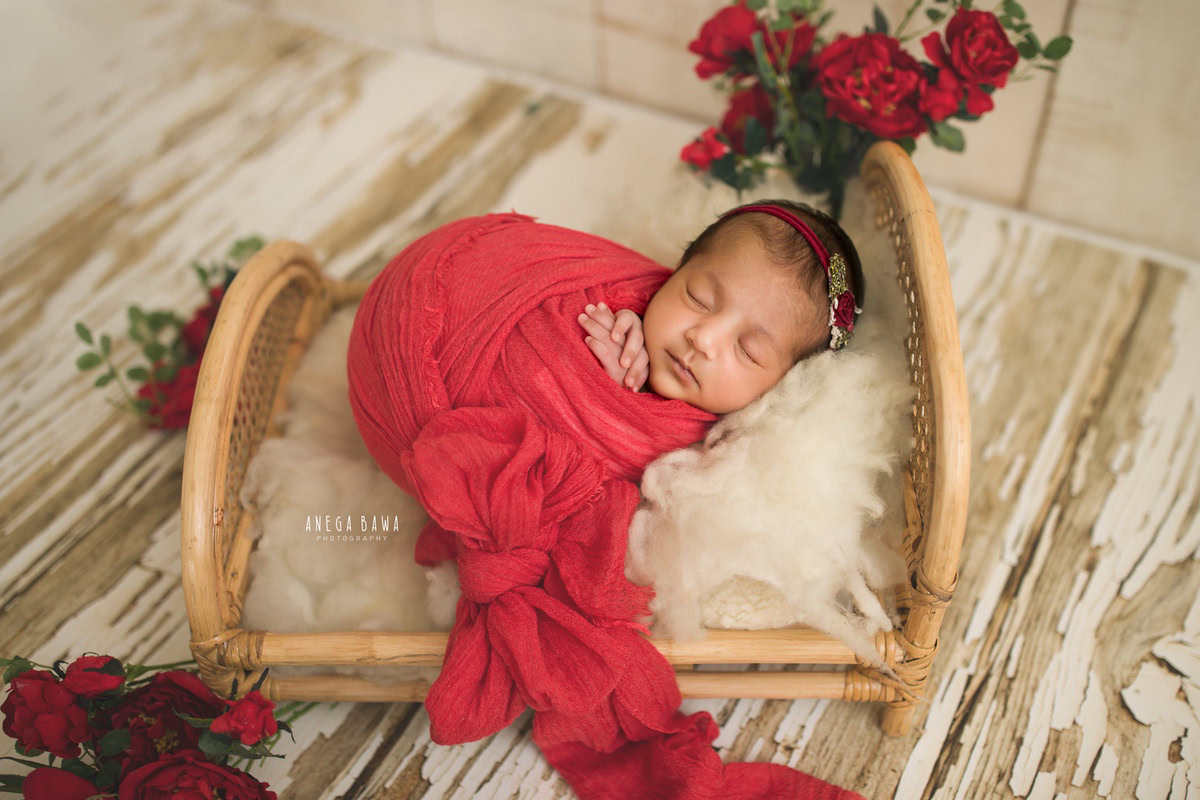 Newborn Photoshoot in Gurgaon: Red-wrapped baby lying in a wooden basket adorned with red flowers, set against a white backdrop. Photographer: Anega Bawa Photography, Delhi Noida.