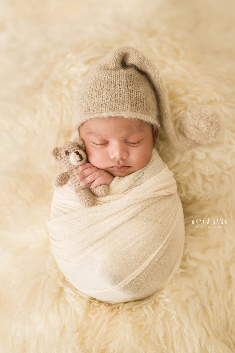 Newborn Photoshoot in Gurgaon: Baby wrapped in white fabric, wearing a brown cap and holding a soft toy, with a beige backdrop. Photographer: Anega Bawa Photography, Delhi Noida.
