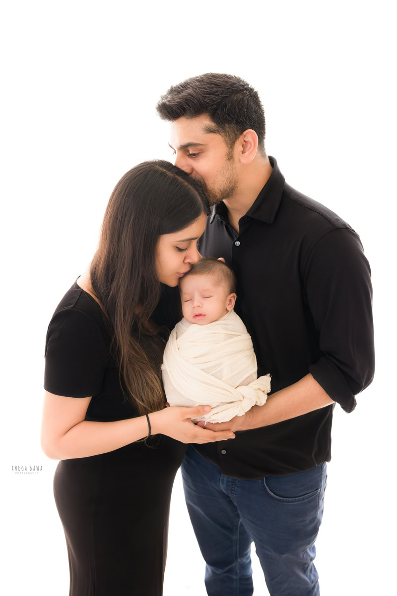 Newborn Photoshoot in Gurgaon: Family with newborn wrapped in white against a white backdrop, wearing black outfits. Photographer: Anega Bawa Photography, Delhi.