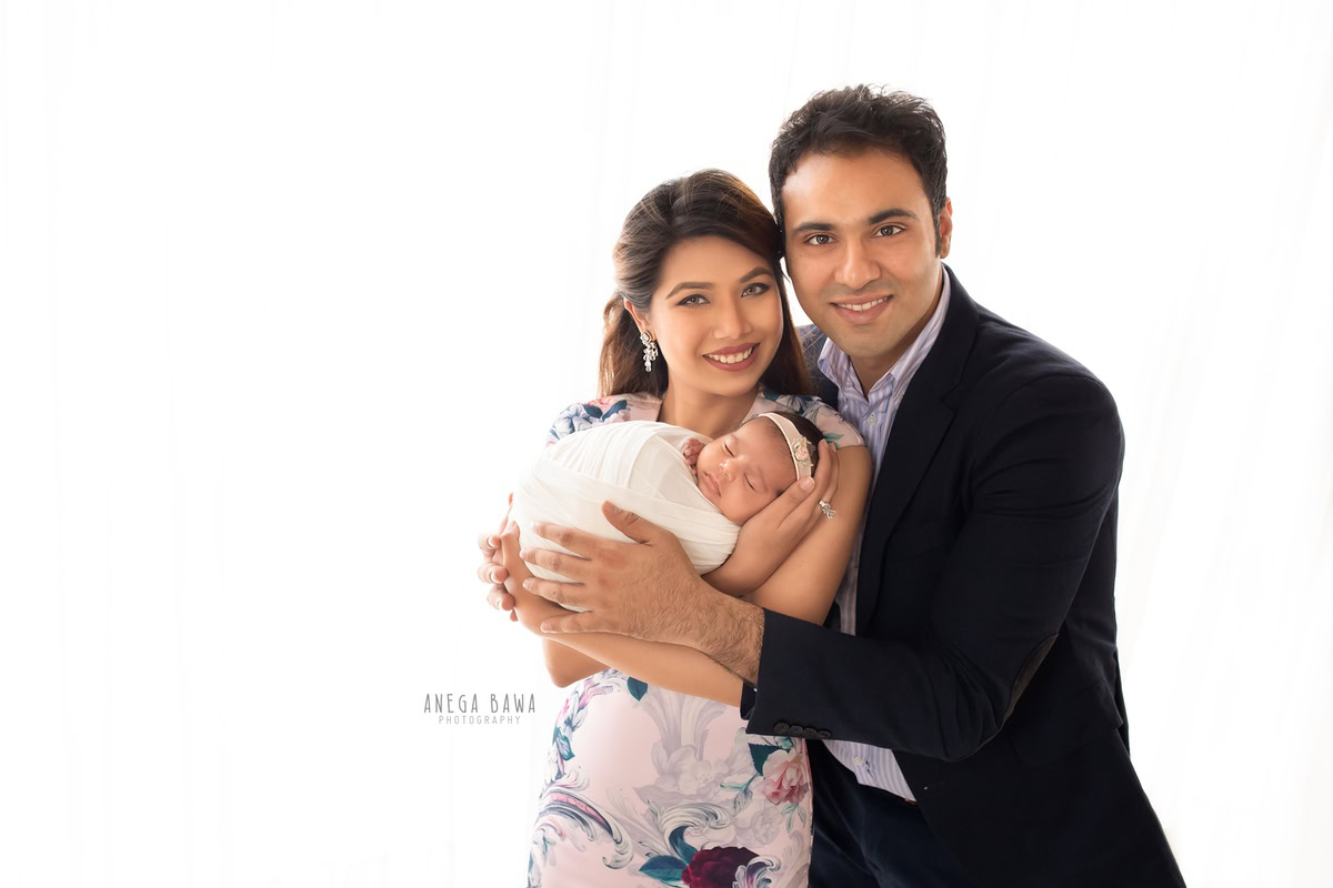 Newborn Photoshoot in Gurgaon: Family with newborn wrapped in white against a white backdrop, wearing floral dresses. Photographer: Anega Bawa Photography, Delhi.