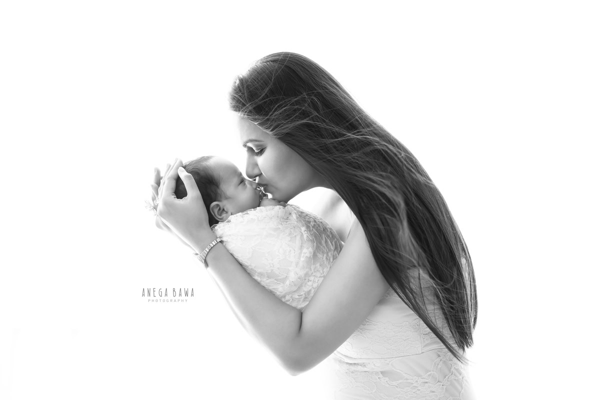 Newborn Photoshoot in Gurgaon: Mother kissing the baby wrapped in white against a white backdrop. Photographer: Anega Bawa Photography, Delhi.