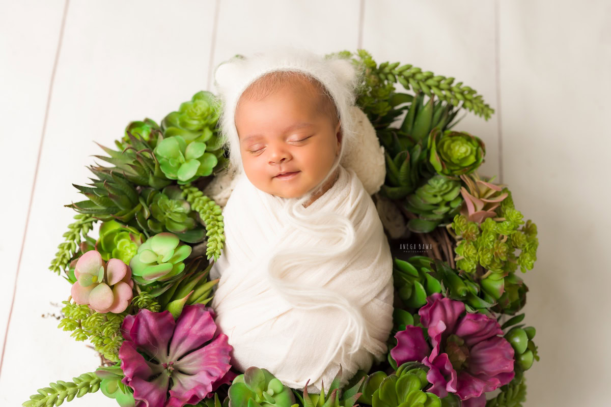 Newborn Photoshoot in Gurgaon: Baby wrapped in white fabric, wearing a white cap, with a cute smile, against a white backdrop. Photographer: Anega Bawa Photography, Delhi Noida.