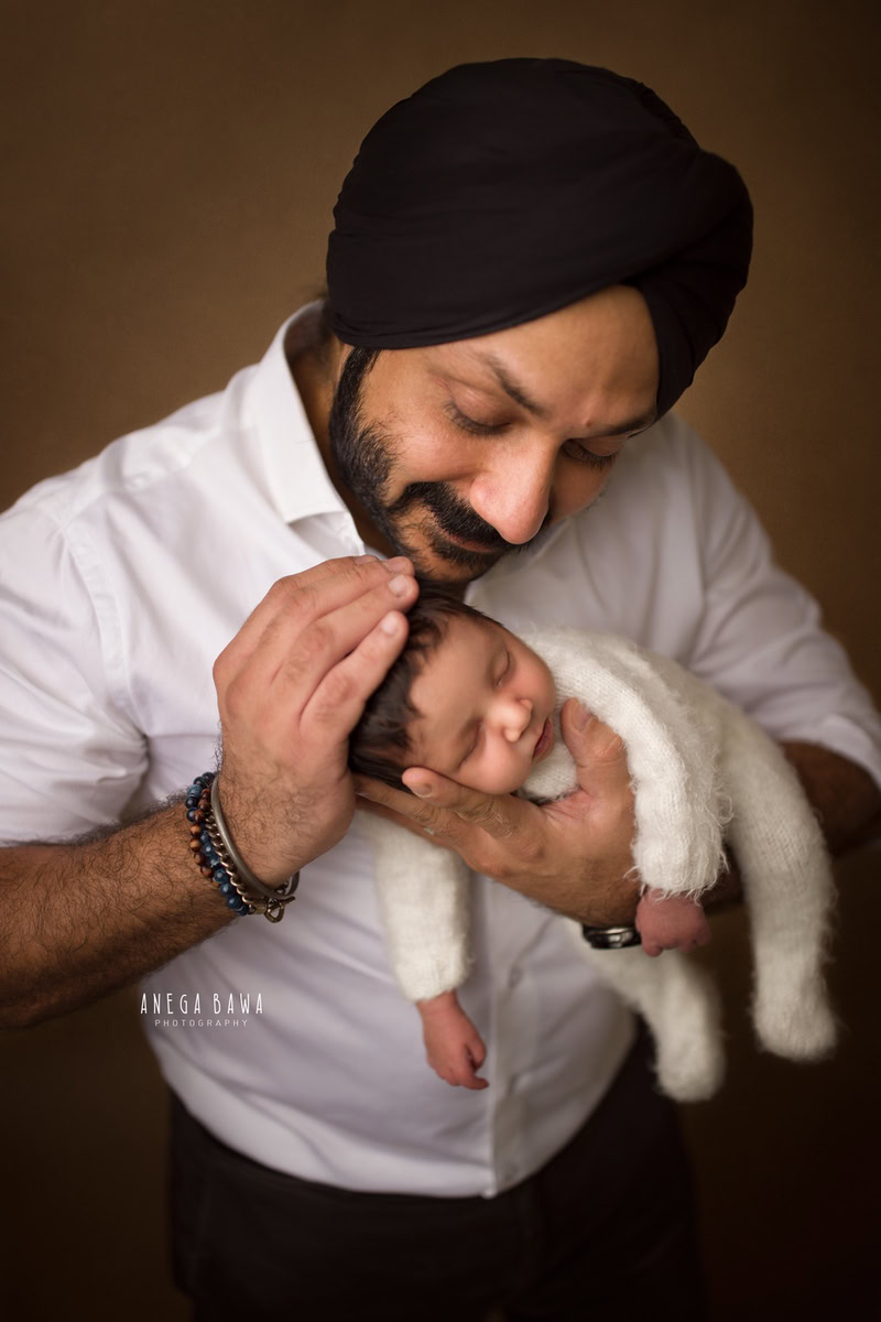 Newborn Photoshoot in Gurgaon: Dad holding newborn in a white baby suit against a brown backdrop. Photographer: Anega Bawa Photography, Delhi.