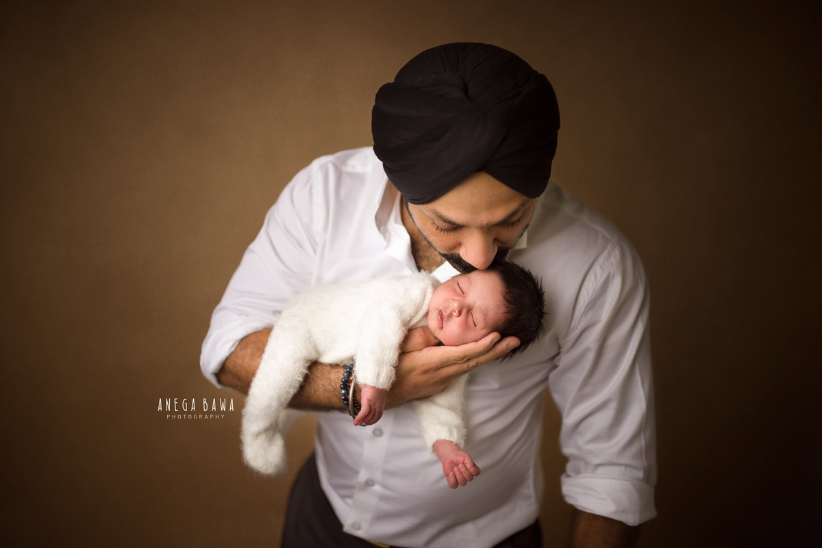 Newborn Photoshoot in Gurgaon: Dad holding newborn in a white baby suit with a Zara pose against a brown backdrop. Photographer: Anega Bawa Photography, Delhi.