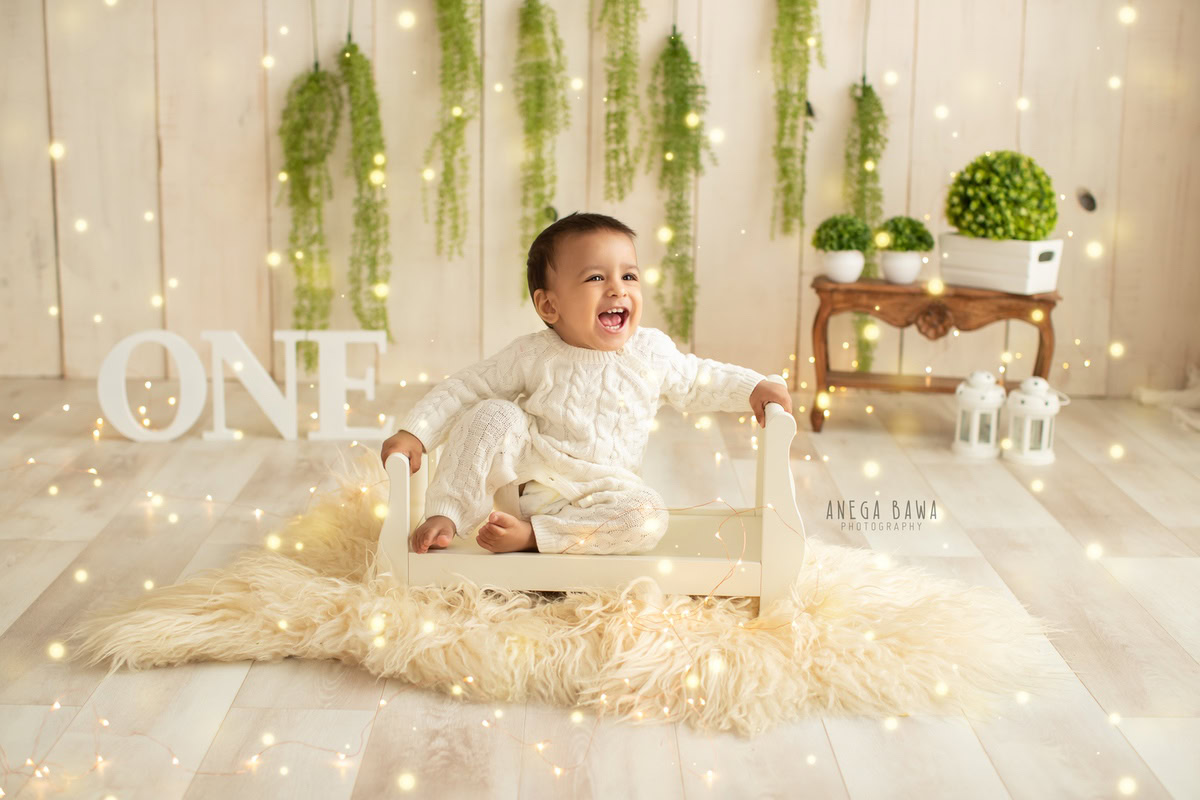 For a sitter photo shoot in Delhi and Gurgaon by Anega Bawa, a 1-year-old boy is captured in a serene and charming setting. Seated in a white cot on a beige rug, surrounded by leafy pots and fairy lights against a beige backdrop, the scene exudes a warm and peaceful ambiance, perfect for commemorating this milestone.