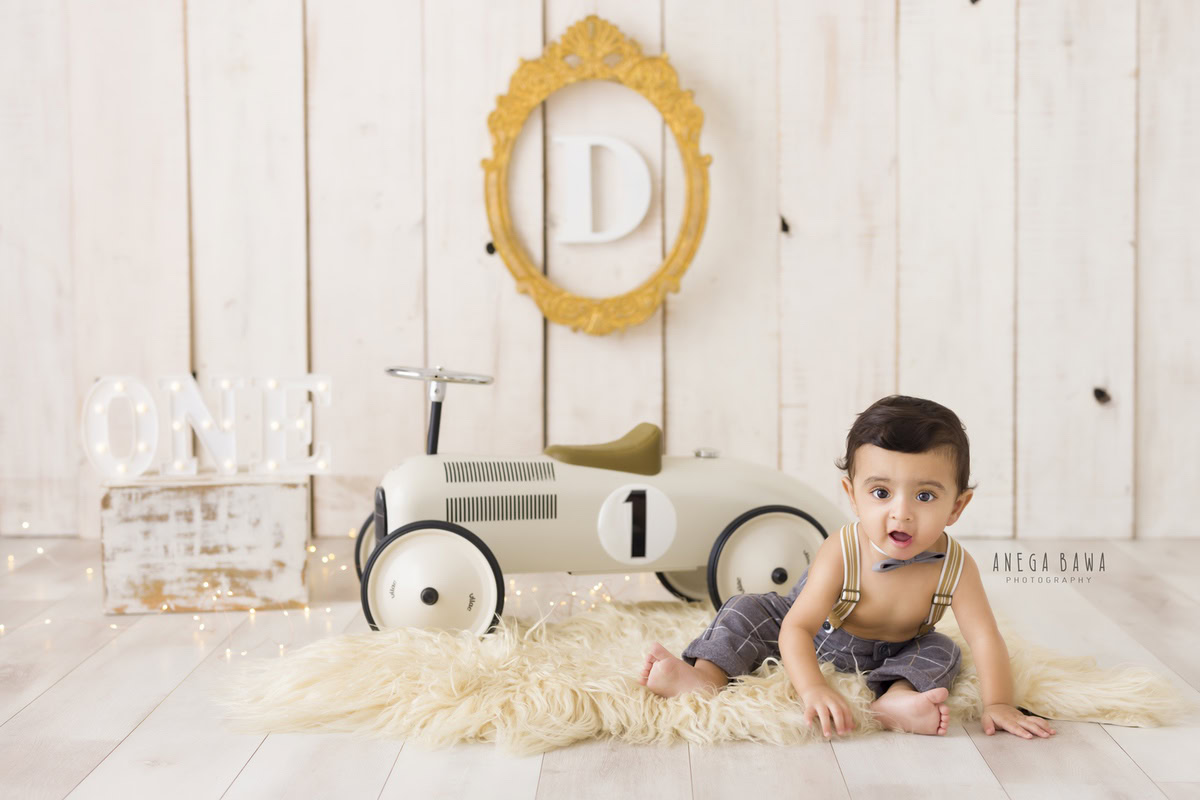 One-year-old boy seated on a beige rug beside a toy scooter with an alphabet frame on the wall against a pristine white backdrop, capturing the essence of his pre-birthday photoshoot in Delhi by Anega Bawa, Gurgaon, Noida.
