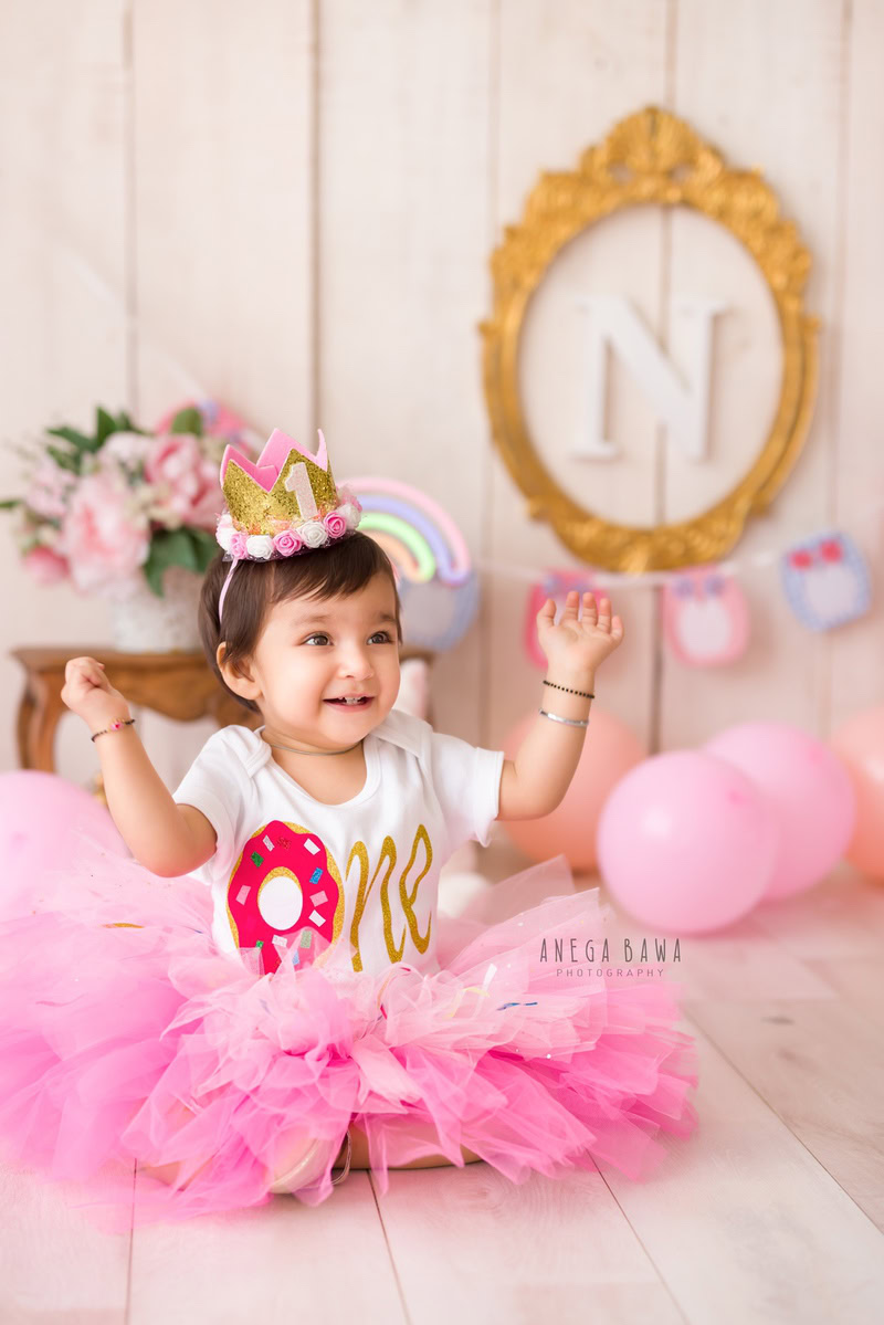 In this delightful sitter photography session by Anega Bawa in Delhi and Gurgaon, a 1-year-old baby girl is dressed in a pink dress and crown. She sits in front of an alphabet frame on the wall, surrounded by nude-colored balloons and flowers in a white vase, creating a whimsical and elegant scene.