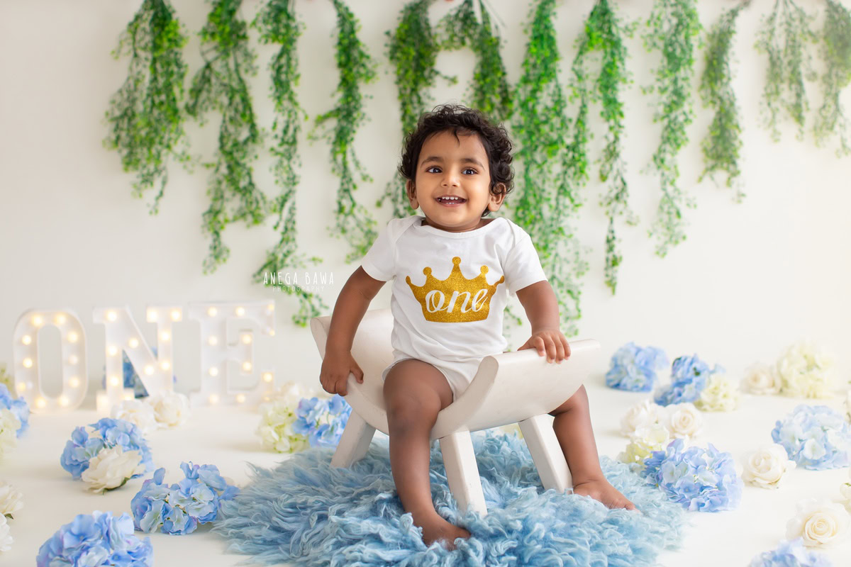 During a sitter photo shoot in Delhi, Gurgaon by Anega Bawa, a 1-year-old boy is captured in his adorable blue babysuit, sitting on a blue rug. The backdrop is tastefully decorated with blue pom-poms and green leafy fringes, set against a crisp white backdrop. This delightful scene beautifully showcases the essence of childhood joy and innocence.