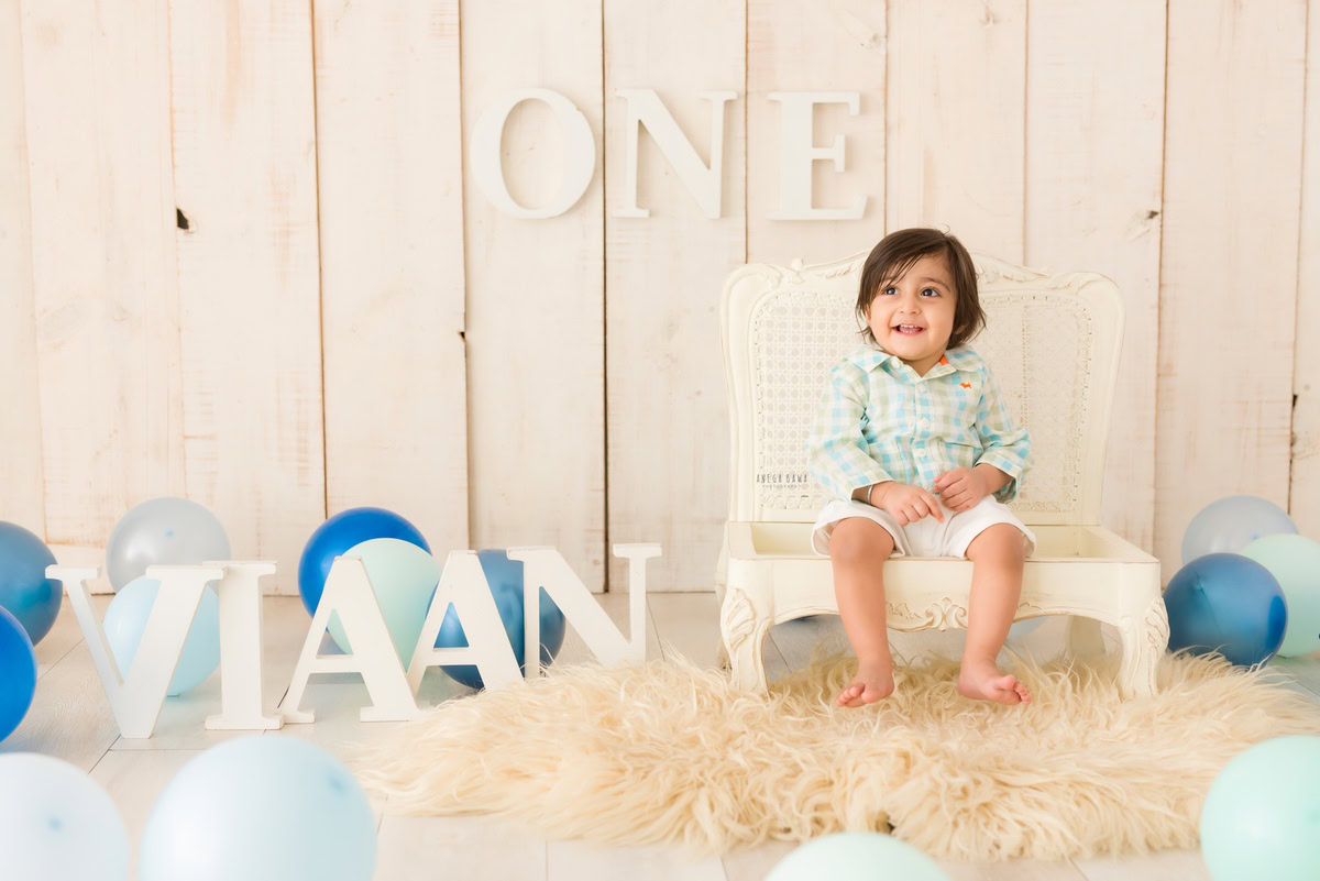 One-year-old boy on a beige rug surrounded by balloons with a name frame on the floor against a beige backdrop, capturing the sweetness of his pre-birthday photoshoot in Delhi by Anega Bawa, Gurgaon, Noida.