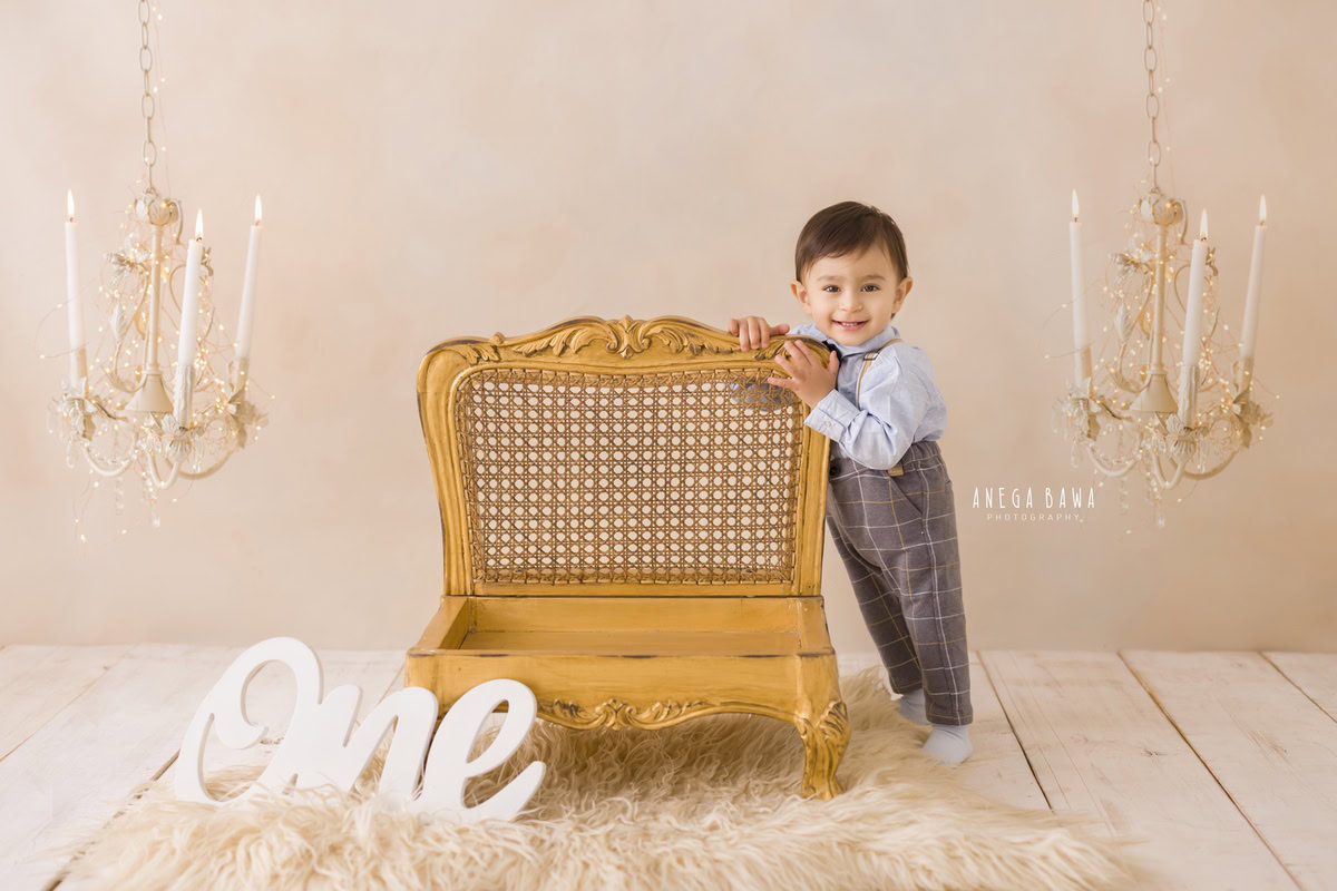 One-year-old boy standing close to a wooden chair with candle stands against a beige backdrop, capturing the charm of his pre-birthday photoshoot in Delhi by Anega Bawa, Gurgaon, Noida.