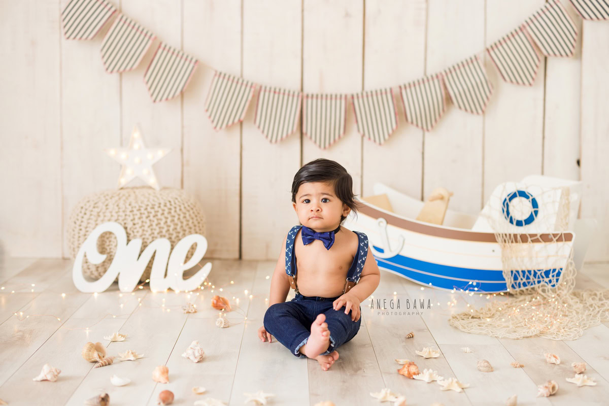 For a sitter photo shoot in Delhi and Gurgaon by Anega Bawa, a 1-year-old boy is charmingly captured with a blue bow, sitting against a beige backdrop. The scene is enhanced with a spaceboat toy in the background and petals scattered on the floor. This whimsical and delightful composition highlights the boy's playful spirit, creating a precious memory to treasure.
