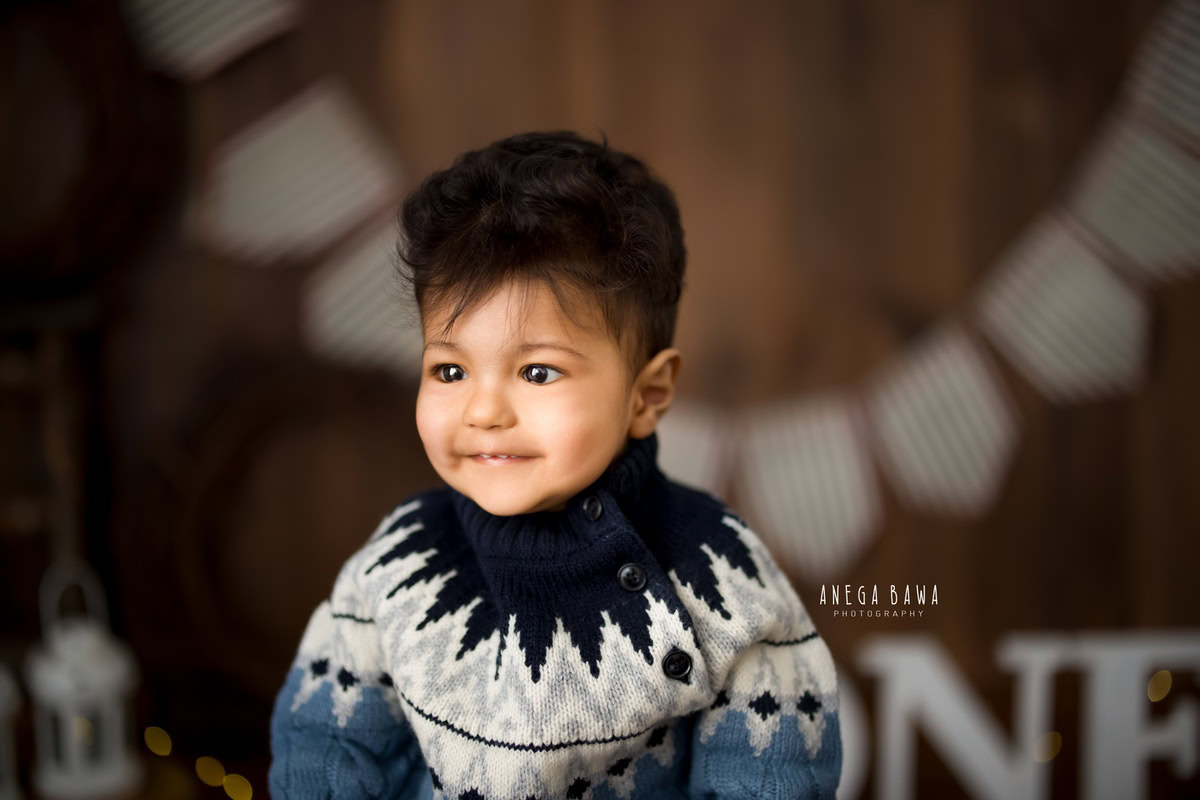 In this charming sitter photography session by Anega Bawa in Delhi and Gurgaon, a 1-year-old boy poses against a brown backdrop. The scene features castles and a fringe on the wall, adding a touch of whimsy and fantasy to the photograph.