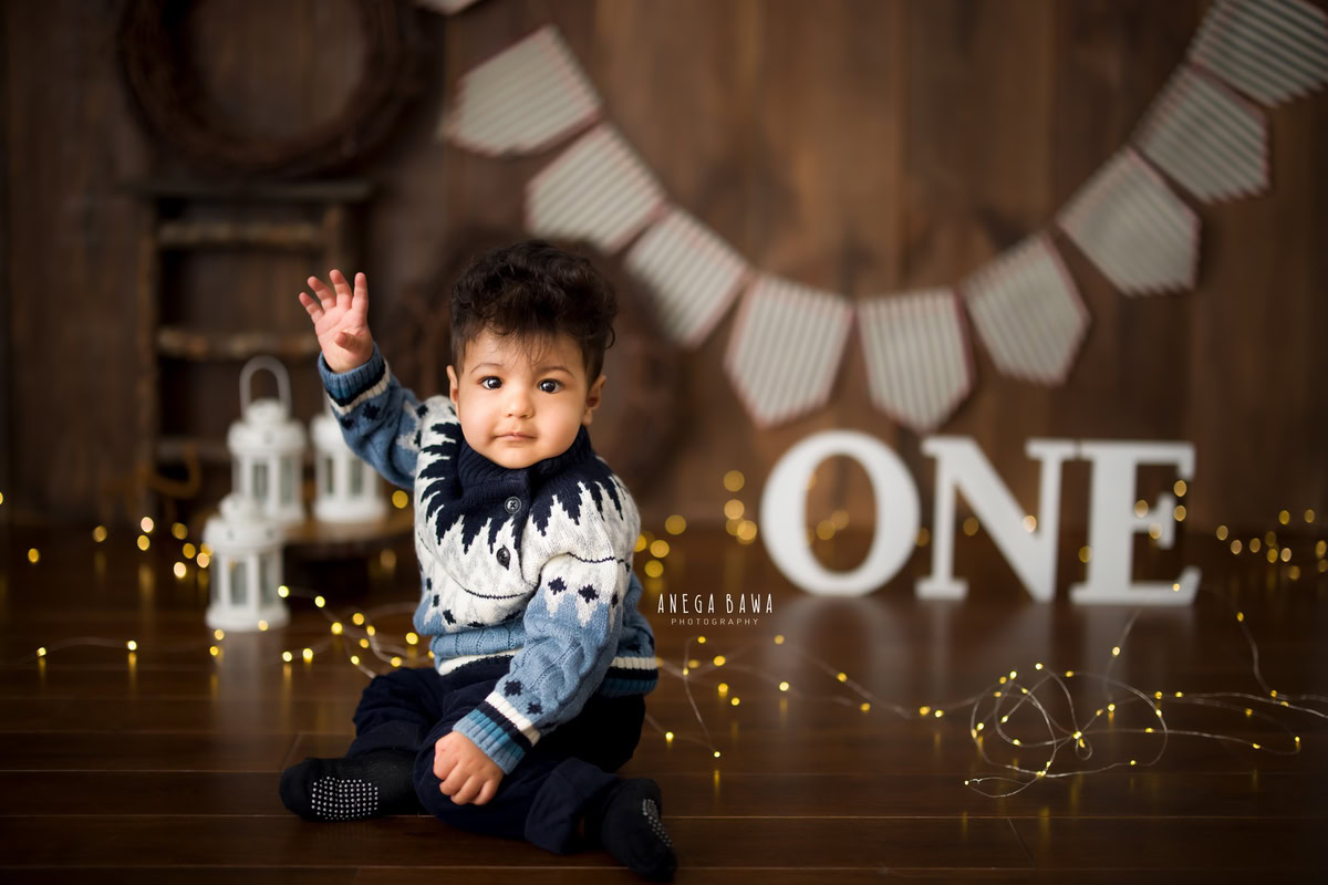 In this delightful sitter photography session by Anega Bawa in Delhi and Gurgaon, a 1-year-old boy poses against a brown backdrop adorned with fairy lights, castles, and a fringe on the wall, creating a magical and enchanting atmosphere.