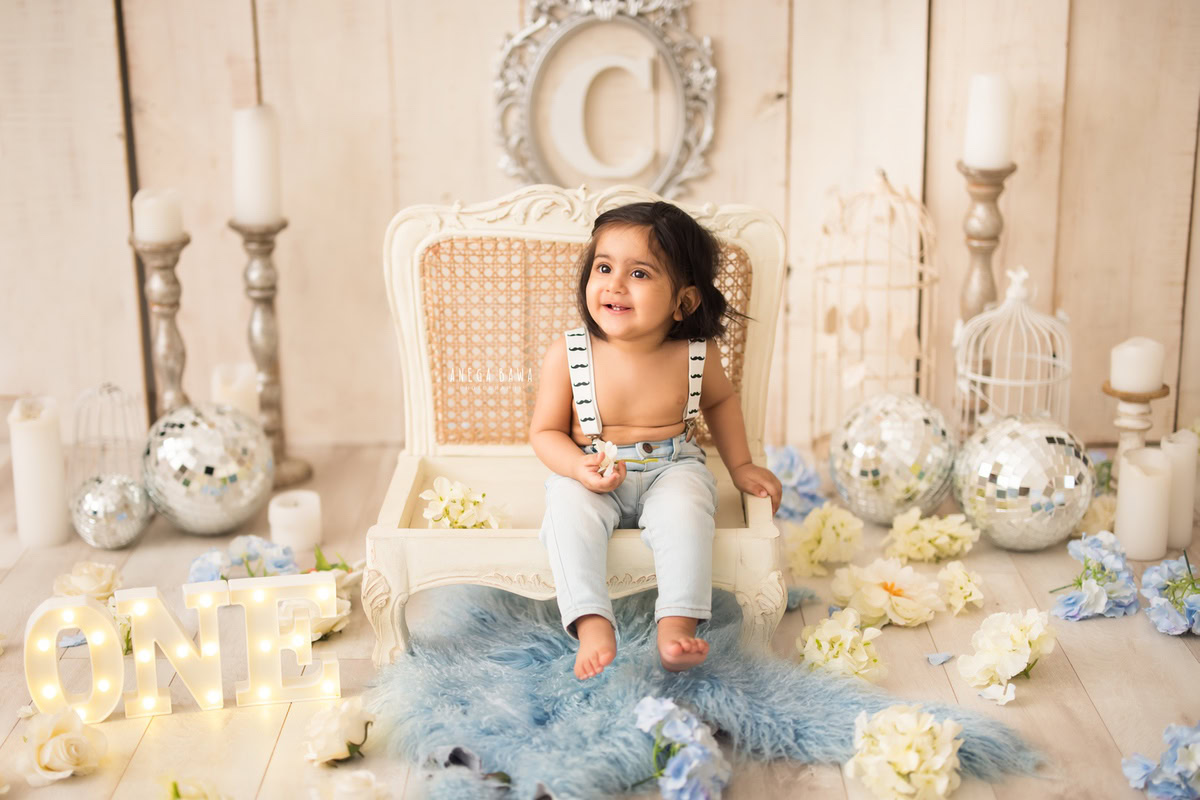 In this enchanting sitter photography session by Anega Bawa in Delhi and Gurgaon, a 1-year-old boy poses with candle stands, disco balls, and castles on a blue rug. The beige backdrop with blue and white flowers adds a charming touch to the scene.