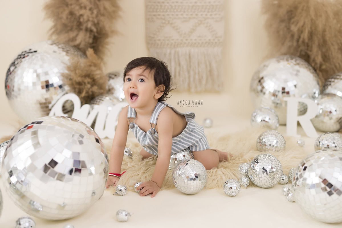 Watch your little explorer venture through a world of wonder, crawling on a soft beige rug amidst shimmering disco balls and lush green bushes. Captured against a pristine white backdrop, this delightful moment is immortalized by Anega Bawa Photography in a captivating sitter photoshoot in Delhi and Gurgaon.