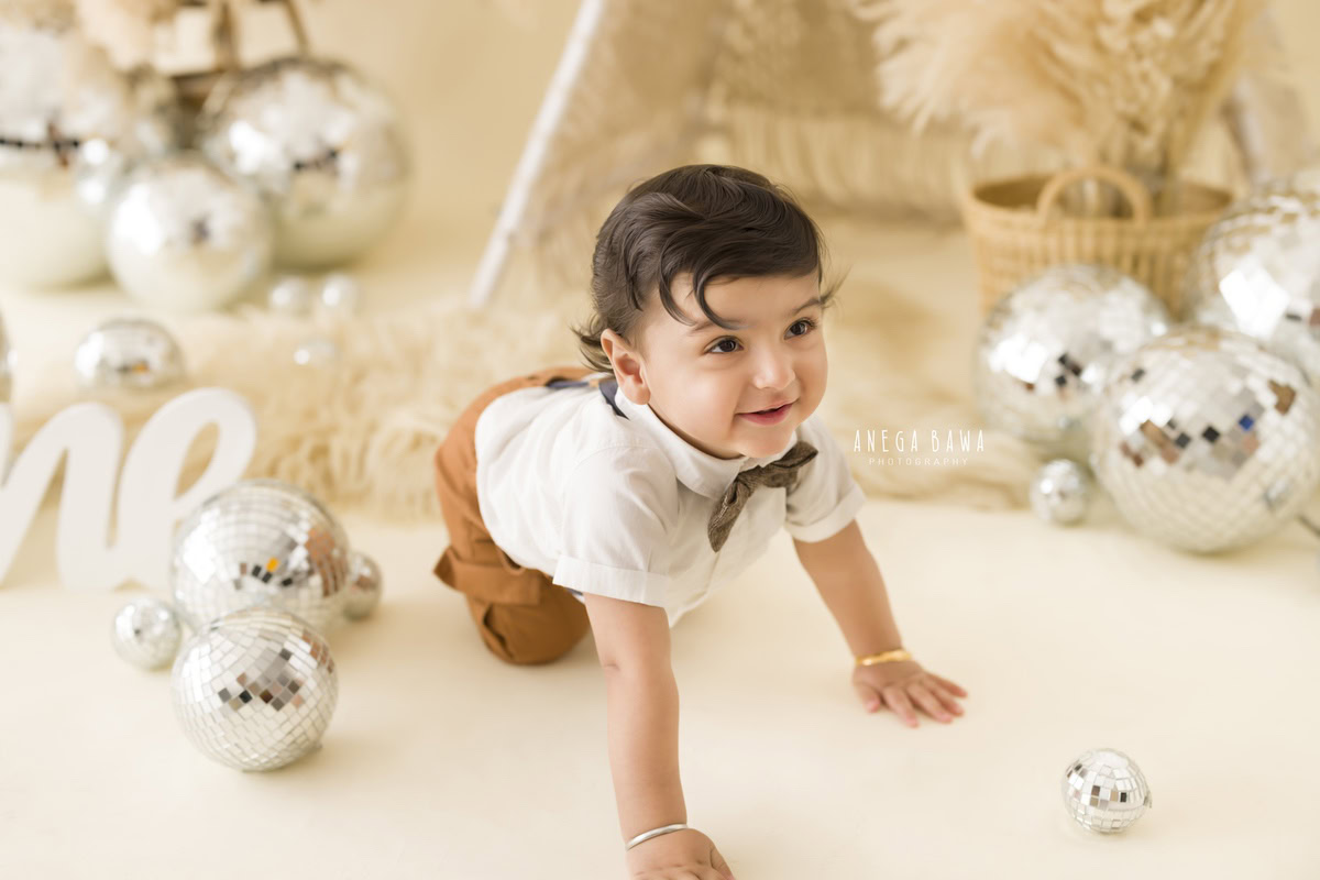 Experience the joyous exploration of your little adventurer as he crawls amidst twinkling disco balls and lush green bushes, all against a soothing beige backdrop. Let Anega Bawa Photography immortalize these precious moments in a delightful sitter photography session in Delhi and Gurgaon.