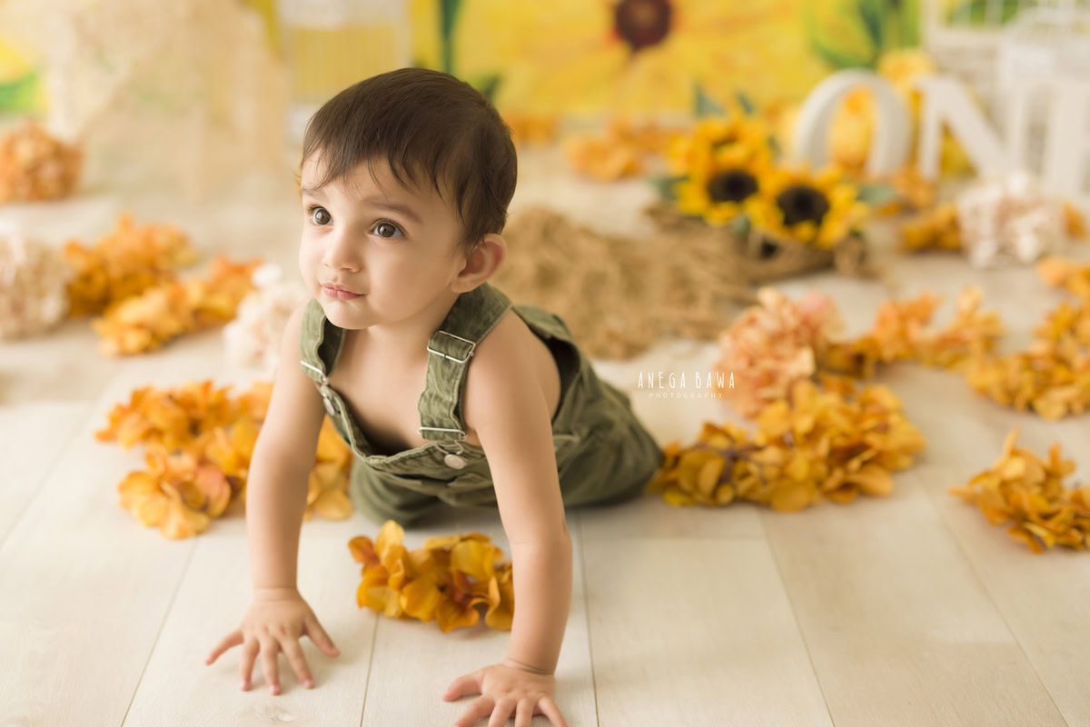 In this sitter birthday photoshoot by Anega Bawa in Delhi, an 8-month-old boy in a blue babysuit and cute cap sits on a blue rug against a cloudy backdrop. This setup highlights his innocence and joy, capturing a precious milestone.